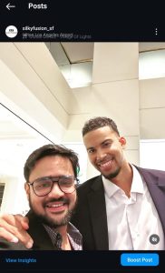 Two smiling individuals are standing together in a well-lit indoor setting. The person on the left, sporting Silky Fusion Hair extensions, wears glasses and a dark blazer over a checkered shirt. The person on the right is clad in a dark jacket with a light shirt. Both appear relaxed and happy.