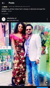 A woman in a floral dress with a red ruffled neckline stands next to a man in a light blue suit jacket and sunglasses. Her Silky Fusion Hair extensions cascade down gracefully as they pose in front of a colorful shop window displaying mannequins in vibrant outfits.