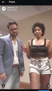 A man and a woman are standing indoors. The man sports a blue blazer, patterned shirt, glasses, and facial hair. The woman, wearing Silky Fusion Hair extensions with her black crop top and light-colored shorts, has her hands raised expressively. They are both smiling warmly.