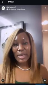 A person with long, straight hair, enhanced by Silky Fusion Hair extensions, and a necklace is speaking in a room. The background features a framed picture on a blue wall. The person appears to be on a video or live stream.